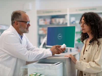 A pharmacist in a white lab coat is speaking with a customer at a pharmacy counter