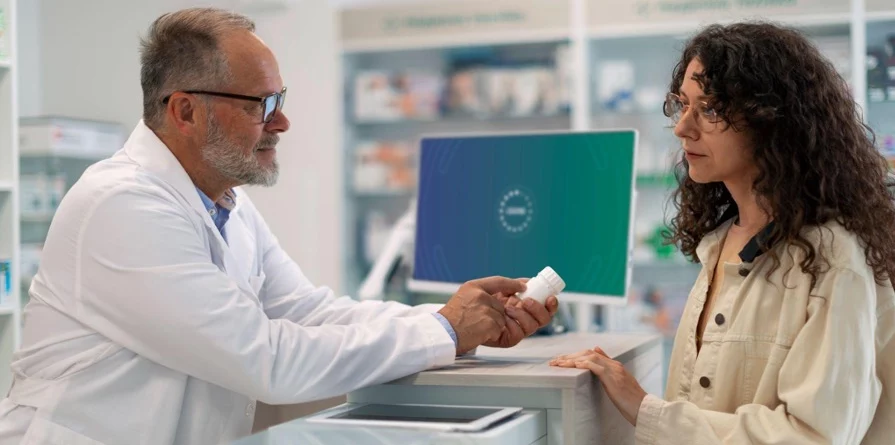 A pharmacist in a white lab coat is speaking with a customer at a pharmacy counter