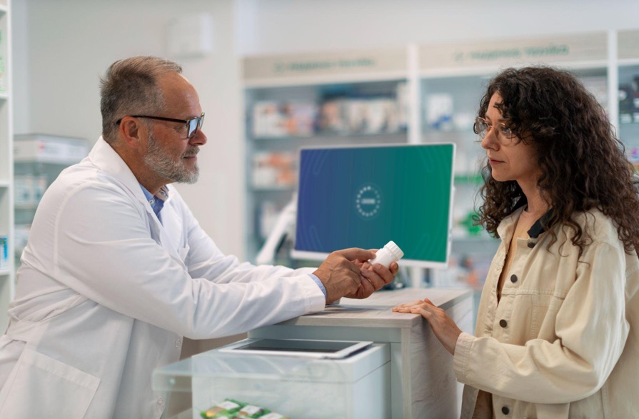 A pharmacist in a white lab coat is speaking with a customer at a pharmacy counter